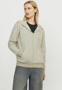 Haut de hoodie zippé vert clair avec une capuche à cordon, des poches avant et des poignets côtelés. Porté sur un tee-shirt noir avec un minimum de détails sur le logo.