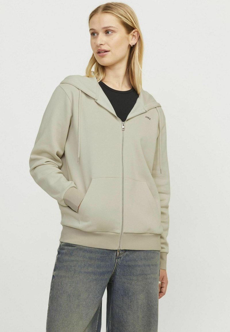 Svetlozelena zip-up kapuca z zategovanjem, sprednjimi žepi in rebrastimi manšetami. Nosi se čez črno majico, ki ima minimalne podrobnosti logotipa.