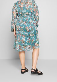 Robe à motif floral en bleu canard avec des manches longues, une ceinture à la taille et une silhouette fluide. Portée avec des sandales plates noires.