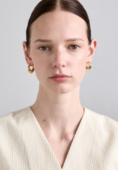 Pendientes de aro de oro con un acabado suave, llevados por una persona de piel clara, que lleva una blusa de tela crema texturizada con un diseño de escote en V.