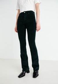 Pantalon en velours côtelé vert foncé, ajusté, avec une fermeture à bouton et une légère évasement en bas. Porté avec des bottines noires à talons blocs.