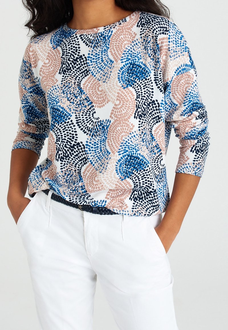 Femme portant une chemise à manches longues avec des motifs abstraits en points bleus, noirs et beiges, un pantalon blanc et une ceinture noire tressée, les mains dans les poches.
