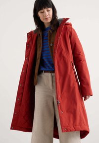 Parka rossa con chiusura a zip e bottoni, caratterizzata da un interno trapuntato e un cappuccio foderato in grigio. Indossata sopra una camicia a righe blu e marrone.
