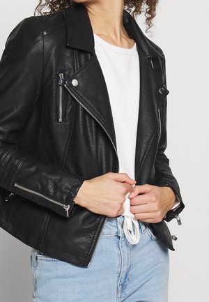Personne portant une veste en cuir noire sur une chemise blanche nouée et un jean bleu clair, tenant le col de la veste avec les deux mains.