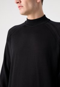 Schwarzer Strickpullover mit hohem geripptem Kragen, der zarte Texturen und Nahtdetails an den Schultern aufweist. Weich und figurbetont.