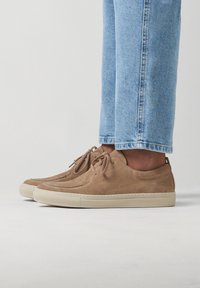 Baskets en daim tan avec des semelles en caoutchouc beige, présentant un design classique à lacets et des détails de couture subtils, portées avec un jean bleu clair.