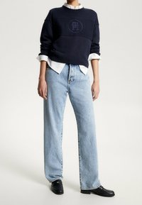 Person, der eine hellblaue, weit geschnittene Jeans, einen marineblauen Pullover über einem weißen Hemd und schwarze Slipper trägt, steht vor einem einfarbigen Hintergrund.
