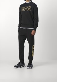 Schwarzes Sweatshirt- und Hosen-Set mit einem metallisch goldenen "VERSACE JEANS COUTURE"-Logo. Das Material ist weich, mit eng anliegenden Bündchen und Seitentaschen.