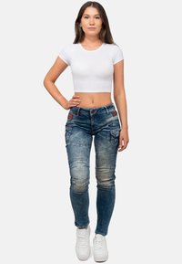 Jeans in denim fitted di colore blu scuro con aree sbiadite, cuciture decorative e tasche laterali; abbinate a un crop top bianco semplice e sneakers bianche.