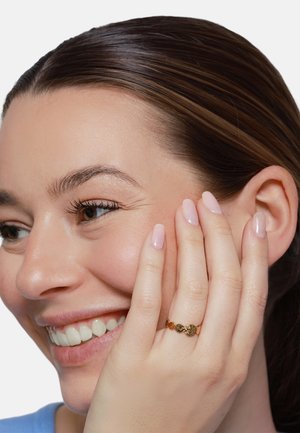 Lächelnde Frau mit zurückgestrichenen braunen Haaren, die die Wange berührt und einen goldenen Ring mit strukturiertem kreisförmigem Design am linken Ringfinger trägt.