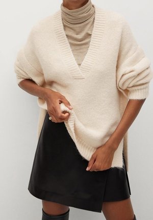 Beżowy, oversize sweter dzianinowy z głębokim dekoltem w serek, prążkowanymi detalami i podwiniętymi rękawami, noszony na jasnobrązowym golfie oraz czarnej skórzanej spódnicy.