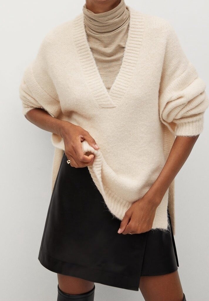 Beżowy, oversize sweter dzianinowy z głębokim dekoltem w serek, prążkowanymi detalami i podwiniętymi rękawami, noszony na jasnobrązowym golfie oraz czarnej skórzanej spódnicy.