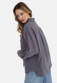 Koszula z długim rękawem, zapinana na guziki, w teksturowanej ciemnoszarej tkaninie z zakładką z tyłu i zaokrąglonym dołem, połączona z jasnoniebieskimi spodniami dżinsowymi.