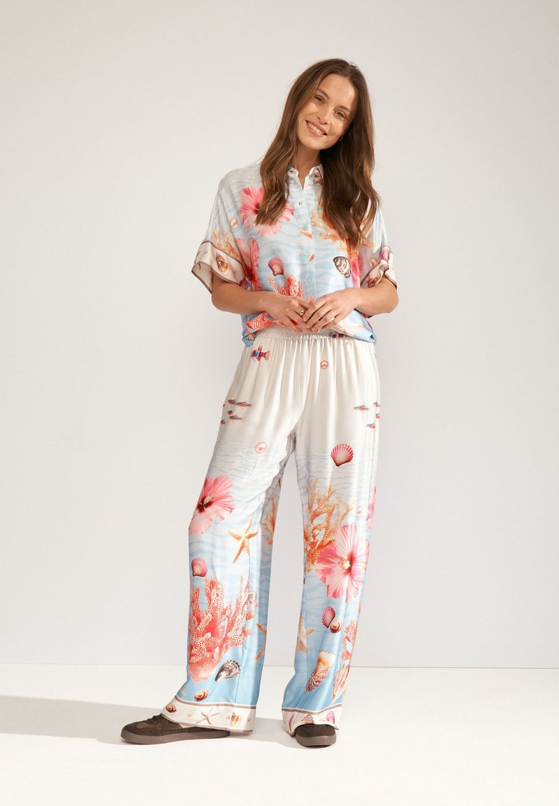 Femme souriante debout portant une chemise assortie bleu et blanc à motifs floraux et coquillages et un pantalon ample avec des chaussures foncées à enfiler.