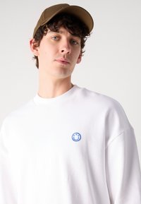 Sudadera blanca de algodón, con un logo de carita sonriente bordado en azul. El modelo lleva una gorra marrón, mostrando un estilo casual.