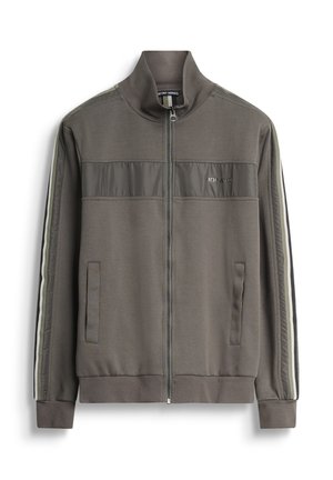 Chaqueta de pista verde aceituna con cierre, dos bolsillos laterales, tejido texturizado y rayas verticales blancas y negras a lo largo de las mangas.