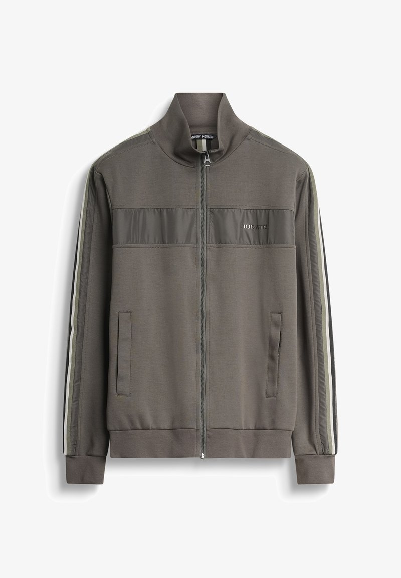 Chaqueta de pista verde aceituna con cierre, dos bolsillos laterales, tejido texturizado y rayas verticales blancas y negras a lo largo de las mangas.