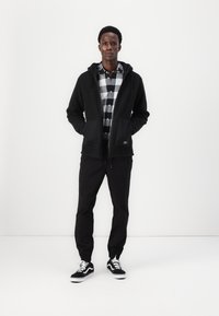Hoodie en polaire noire avec fermeture éclair, doté de deux poches avant. Assorti avec une chemise à carreaux noir et blanc et un pantalon noir. Baskets noires.