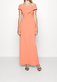Off-the-shoulder coral maxi dress made of smooth fabric with a twisted bodice design and a fitted silhouette.  High slit reveals black heeled sandals.