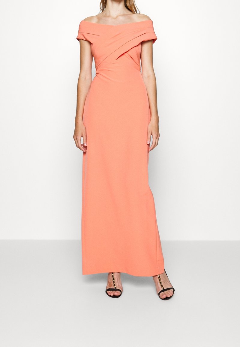 Off-the-shoulder coral maxi dress made of smooth fabric with a twisted bodice design and a fitted silhouette. High slit reveals black heeled sandals.