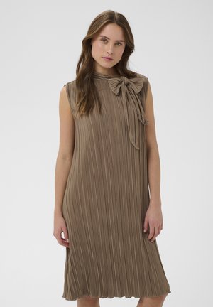 Młoda kobieta o długich włosach ubrana w bezrękawnikową, plisowaną taupe sukienkę z kokardą zawiązaną przy dekolcie, stojąca na jednolitym tle.