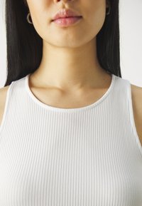 Weißes, geripptes Tanktop mit rundem Ausschnitt, figurbetontem Design und weicher Textur. Das Modell trägt Creolen und hat glatte Haut.