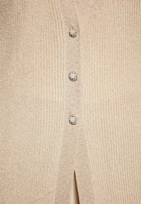 Cardigan tricotat bej, prezentând patru nasturi decorativi cu accente asemănătoare cristalelor și o fanta verticală la tiv.
