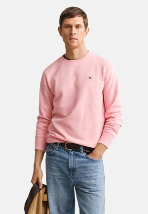 Muž nosi svetloružovú mikinu s dlhým rukávom a modré rifle, drží v jednej ruke hnedú bundu, s neutrálnym výrazom.