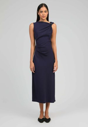 Femme portant une robe midi bleu marine sans manches avec un col asymétrique et des ballerines noires à bout pointu, debout devant un fond gris uni.