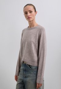 Jonge vrouw met slicked-back haar, gekleed in een licht mauve gebreide trui en blauwe jeans, staande tegen een effen lichtgrijze achtergrond.