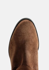 Vârful unei botine din suede maro, cu marginea talpii negre, pe un fundal simplu de gri deschis, văzut din sus.