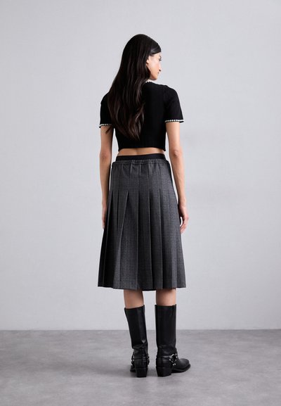 Schwarzer, croppter Pullover mit kurzen Ärmeln, kombiniert mit einem grauen, gefalteten Midikleid. Das Model trägt knielange schwarze Stiefel. Glatte Texturen, karierte Muster.