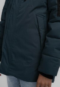 Mörk turkos puffjacka med en framficka, tjock vaddering och svarta tygaccenter på axelområdet samt ribbade muddar.