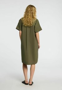 Olive green, sukienka do kolan z krótkimi rękawami, luźny fason i plisowany tył. Noszona z czarnymi, wsuwanymi butami.