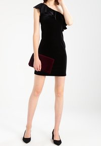 Černé sametové šaty na jedno rameno s volánkovým detailem, doplněné bordó listovou kabelkou a černými podpatky. Hladká textura, přiléhavý střih.