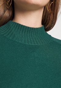 Un maglione verde lavorato a maglia presenta un collo alto a coste e una texture morbida, con un'inquadratura ravvicinata dell'area del collo.