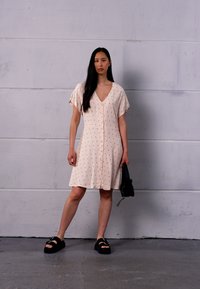 Vestido curto rosa claro com decote em V, mangas curtas, delicada padrão de bolinhas pretas, fecho de botão e um corte solto. Usado com sandálias pretas.