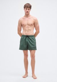 Zaļas peldbikses no viegla auduma ar auklu jostasvietā un sānu kabatām, priekšpusē ar nelielu melnu logotipu.