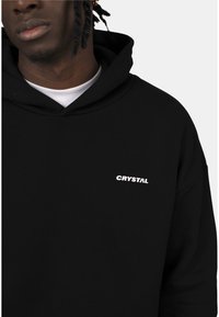 Felpa nera con cappuccio, caratterizzata da un logo bianco "CRYSTAL" sul petto, realizzata in morbido cotone e dal taglio comodo.