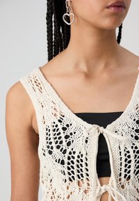 Krótki sweter na drutach w kolorze ecru z ażurowym wzorem, z wiązaniem z przodu i czarną warstwą wewnętrzną. Srebrny kolczyk w kształcie serca dodaje detalu.