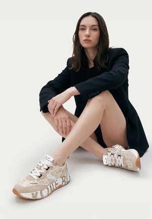 Femme aux longs cheveux foncés portant un blazer noir et des baskets beiges à motifs, assise par terre, les jambes croisées.