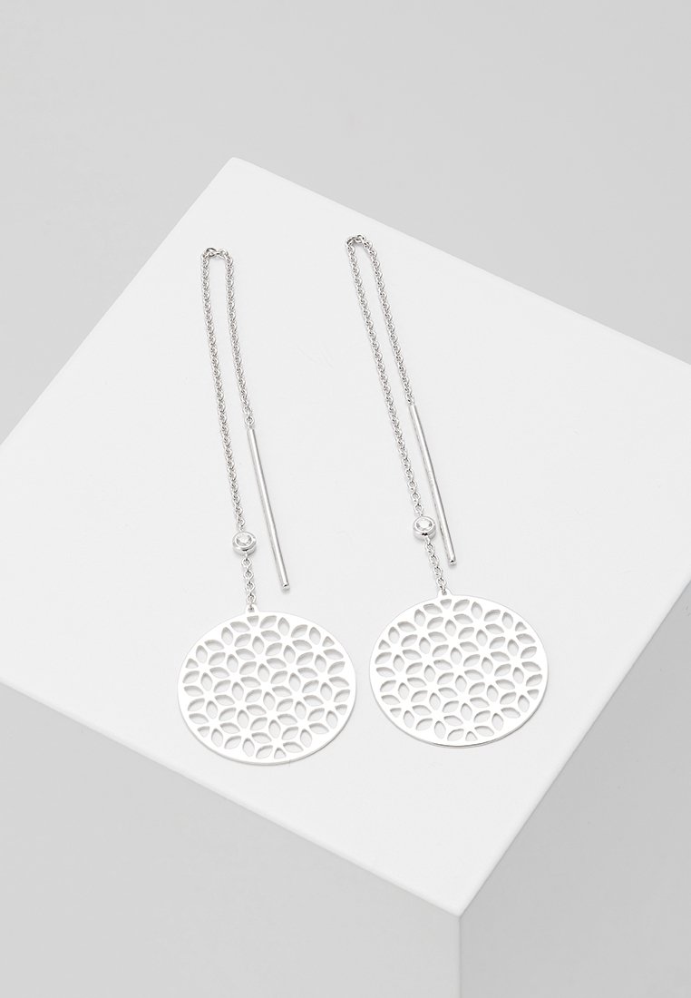 Boucles d'oreilles en fil d'argent avec pendentifs ronds à motif floral ajouré sur une surface géométrique blanche.