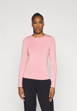 Long-sleeve pink top made from soft fabric, featuring a round neckline and a subtle logo accent near the hem. Paired with black pants.