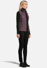 Purple puffer vest with a high collar, visible stitching, and front zipper, worn over a black long-sleeve top and paired with black pants and boots.