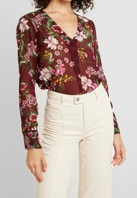 Kobieta w bordowej, kwiecistej bluzce z długim rękawem, włożonej w wysokie, kremowe spodnie, stojąca z rękami swobodnie opadającymi wzdłuż ciała.