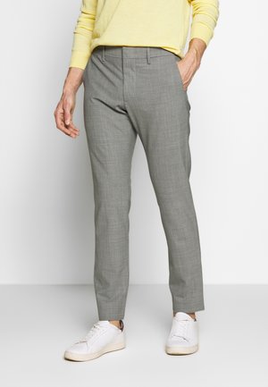 Man draagt grijze slim-fit broek, witte sneakers en een geel shirt met lange mouwen, staat met één hand in zijn zak.