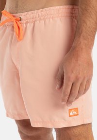 Oranžové plavkové šortky so zebrovaným vzorom, elastickým pásom a šnúrkou na stiahnutie. Obsahujú malú nášivku s logom v oranžovej farbe na spodnej strane.