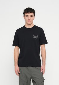 Črna bombažna majica s kratkimi rokavi in okrolim ovratnikom; na levi strani prsnega koša se nahaja subtilen siv logotip.