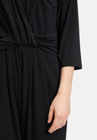Tuta nera con design a vita annodata, maniche corte e tessuto liscio e elasticizzato. Una mano è mostrata posata sul capo.