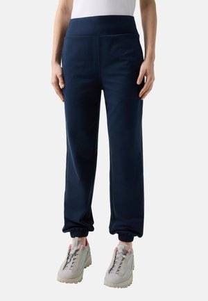 Person trägt dunkelblaue Jogginghosen mit elastischem Bund und hellgraue Turnschuhe mit pinken Akzenten und steht vor einem schlichten Hintergrund.